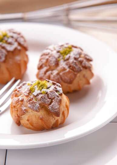 Homemade cream puffs in Lahore topped with powdered sugar and crushed pistachio served on white plate.